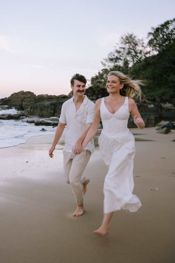 Playful couple running along Coolum Beach during engagement shoot