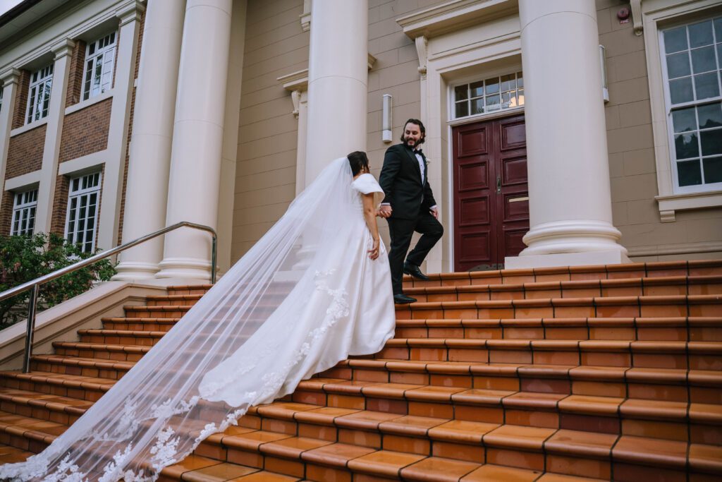 documentary style photo of couple walking up stairs with bride's cathedral veiling trailing behind 