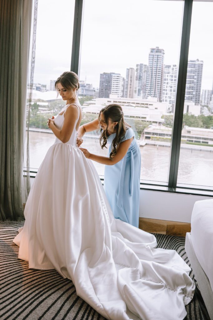 maid of honour doing bride's dress up in front of window with city views