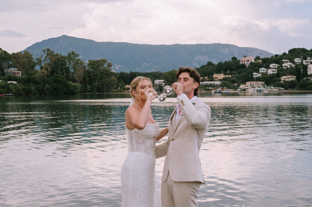candid shot of couple drinking champagne after wedding ceremony with gouvia bay and mountains in background