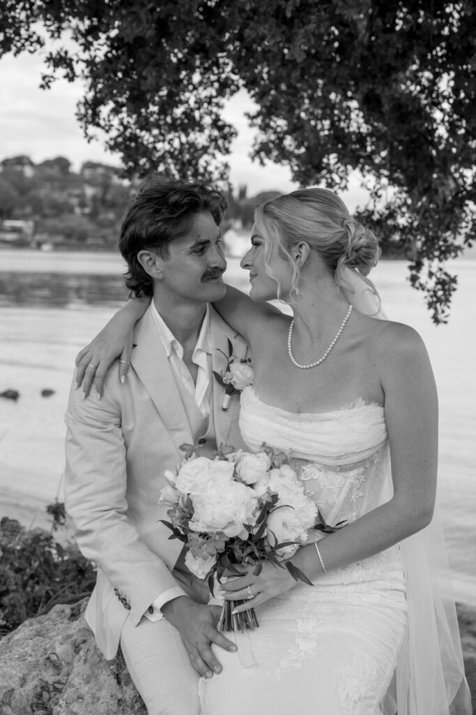 candid black and white shot of bride sitting on groom's lap while holding bridal bouquet