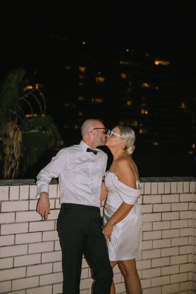editorial style shot of couple kissing while wearing sunglasses and leaning against bricks